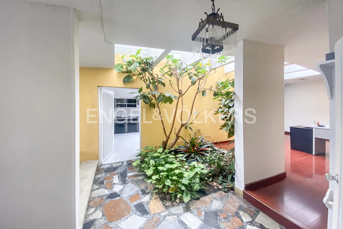 Indoor courtyard with stone floor, plants, and a tree. A white door is open to the outside. A chandelier hangs from the ceiling.