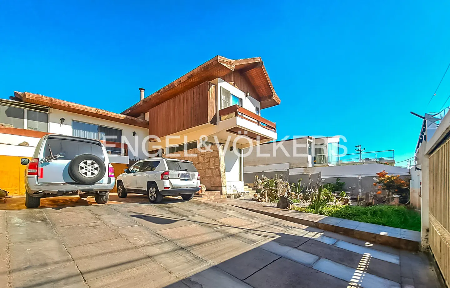 Exterior view of a two-story house with a wood and stone facade, a driveway, and two parked cars under a clear blue sky.