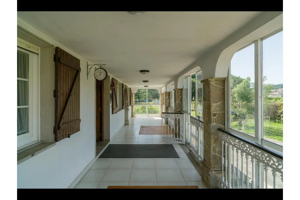 Covered porch with white walls, tile floor, and stone pillars. Windows overlook a green landscape. Shutters and a clock adorn the wall.