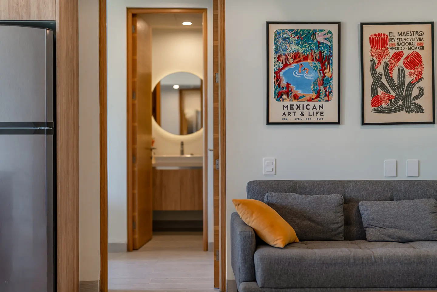 Interior view of a living space with a gray sofa, yellow pillow, and framed art. A bathroom is visible through an open door.