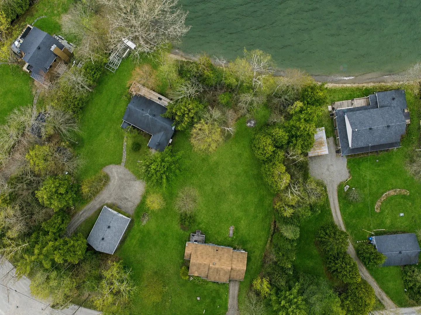 Aerial view of several houses with dark roofs on green lawns near a body of water.