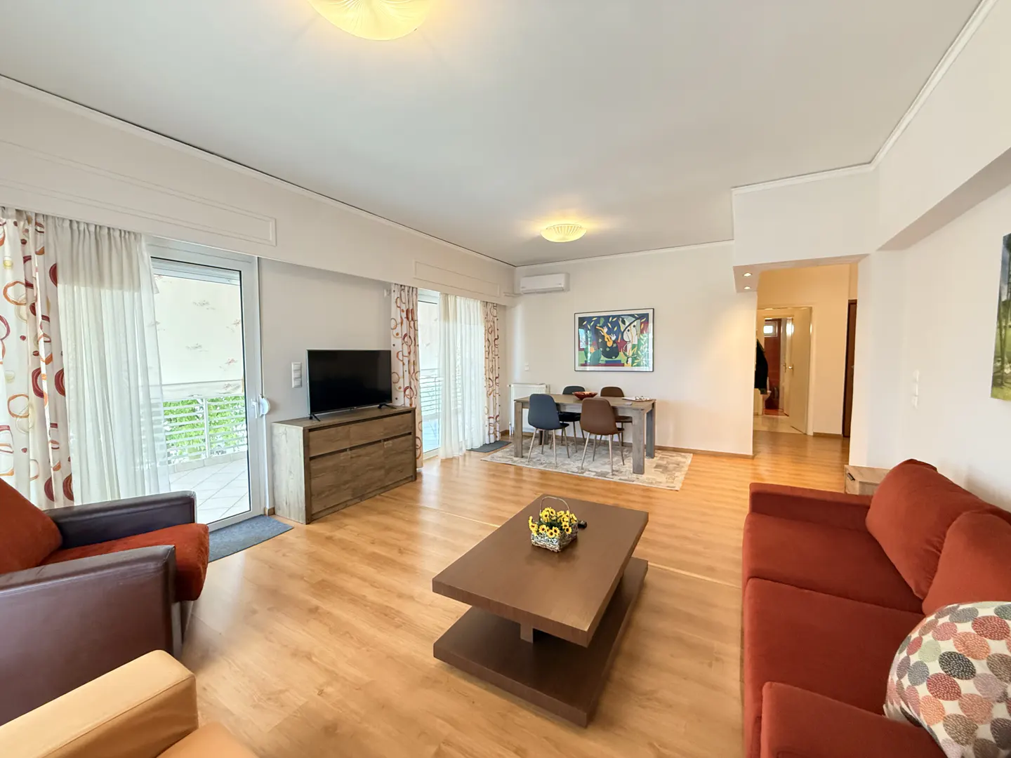 Bright living room with wood floors, a red sofa, a brown coffee table, and a dining table with chairs. Curtains frame a balcony door.