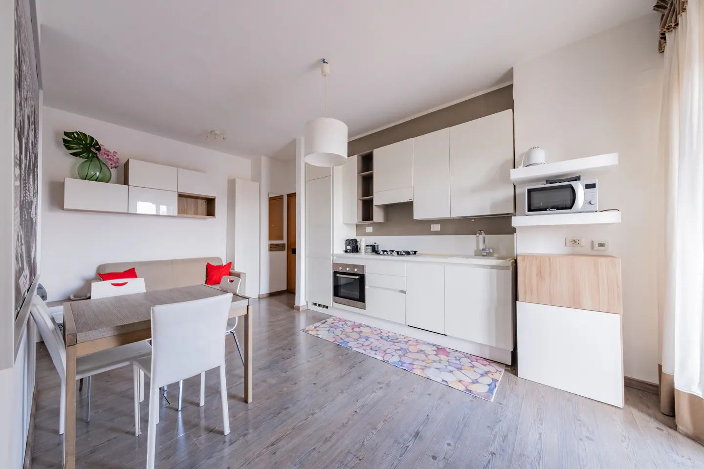 Bright, modern apartment interior with white cabinets, dining table, and gray wood floors. A colorful rug adds a touch of vibrancy.