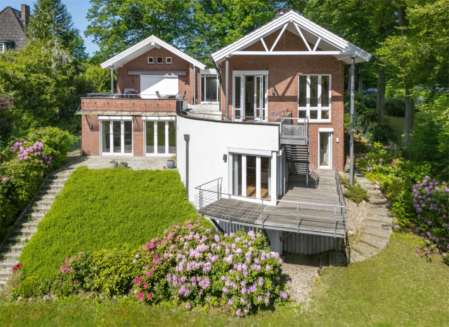Modern brick home with a curved white wall, wooden deck, and lush garden with blooming pink rhododendrons.