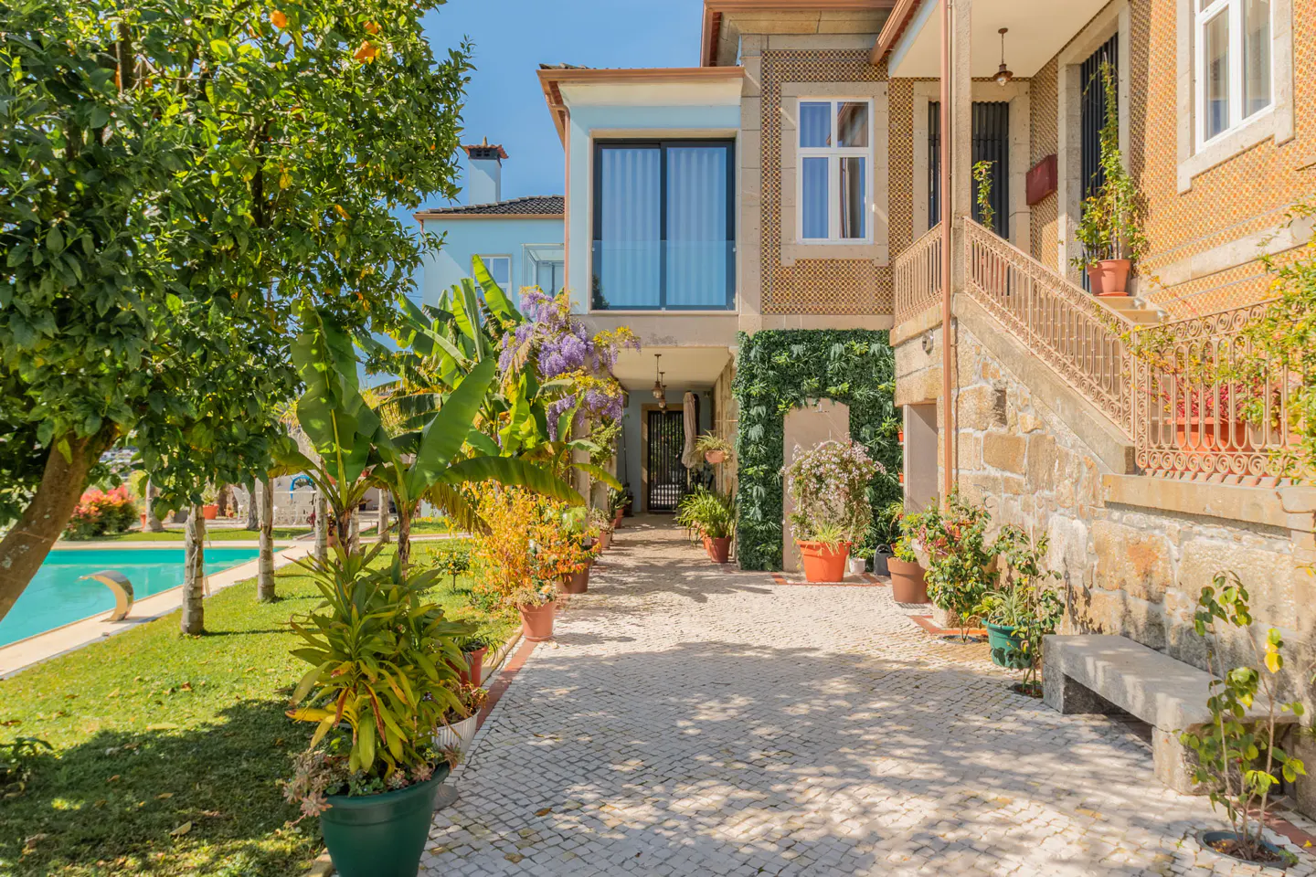 Exterior view of a stone house with a tiled walkway lined with potted plants, leading to a blue pool and lush greenery.