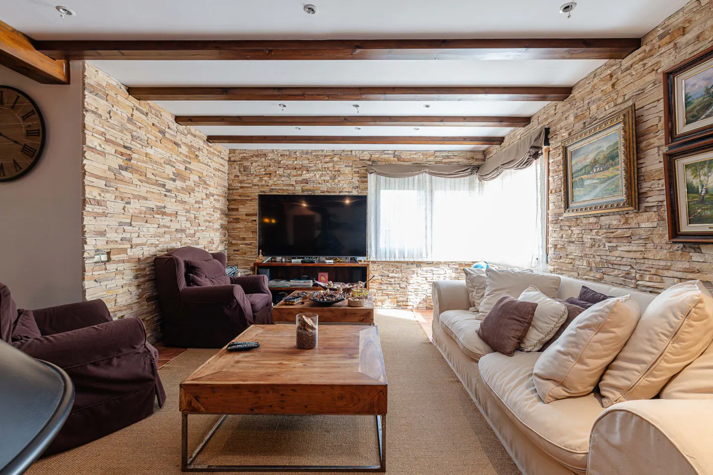Living room with stone walls, wooden beams, and neutral furniture. A large TV sits above a wooden console. Paintings hang on the wall.