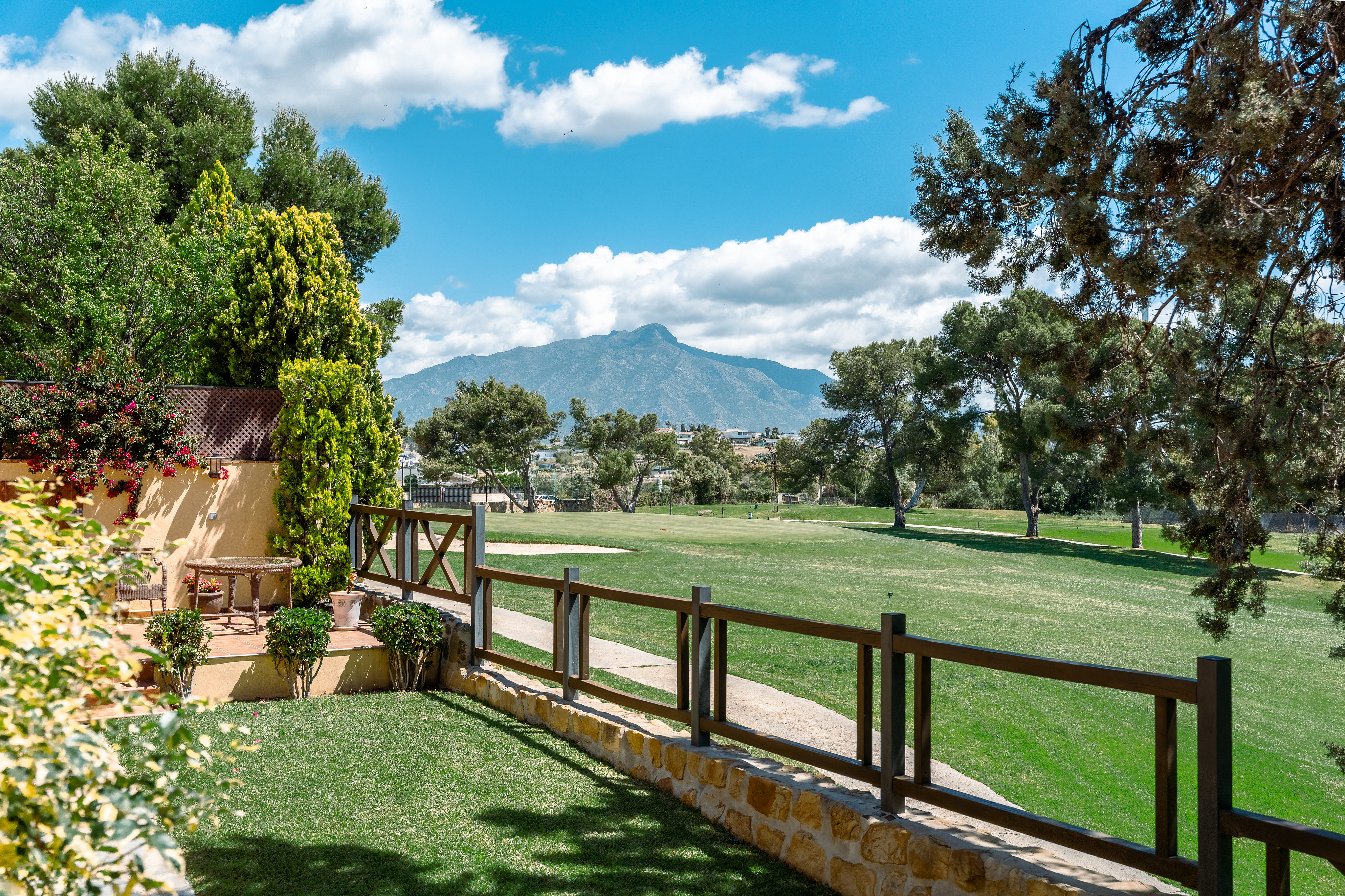 View of a green golf course from a patio with a wooden fence and a mountain in the background under a blue sky.