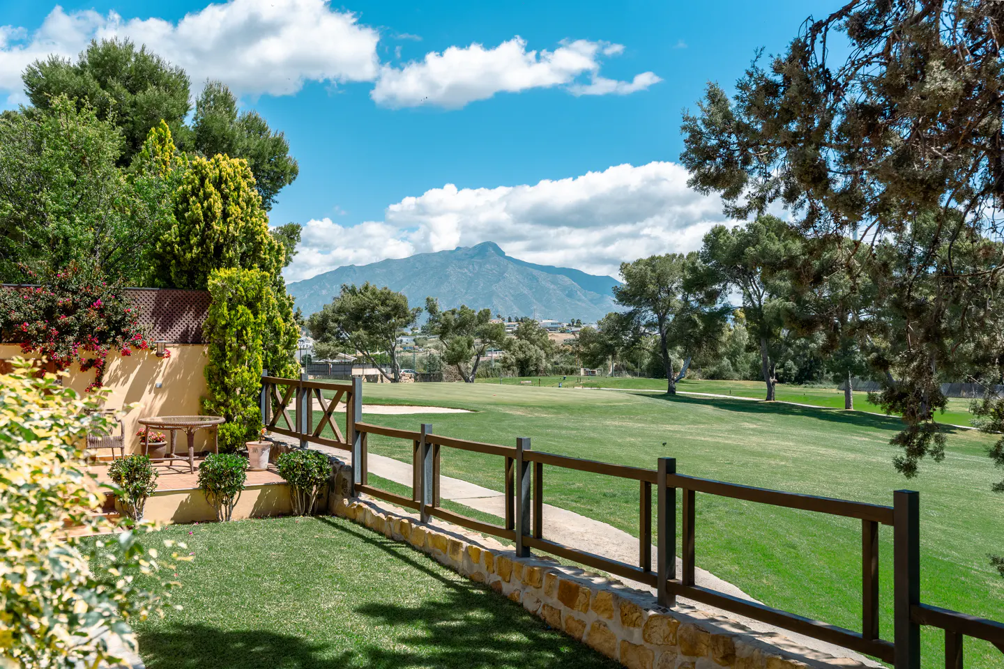 View of a green golf course from a patio with a wooden fence and a mountain in the background under a blue sky.