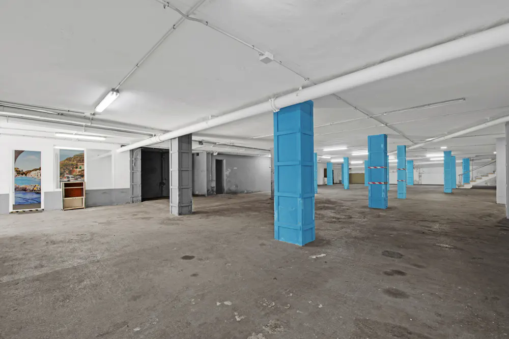 Empty parking garage with gray floor, white ceiling, and blue support columns. Two wall posters are visible on the left.