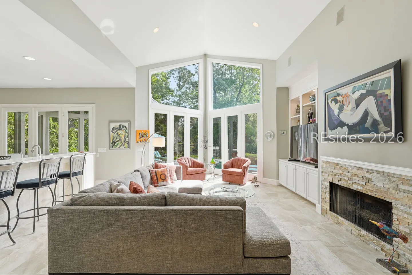 Bright living room with a gray sectional sofa, two pink armchairs, and a stone fireplace with a painting above. Large windows overlook a green yard.