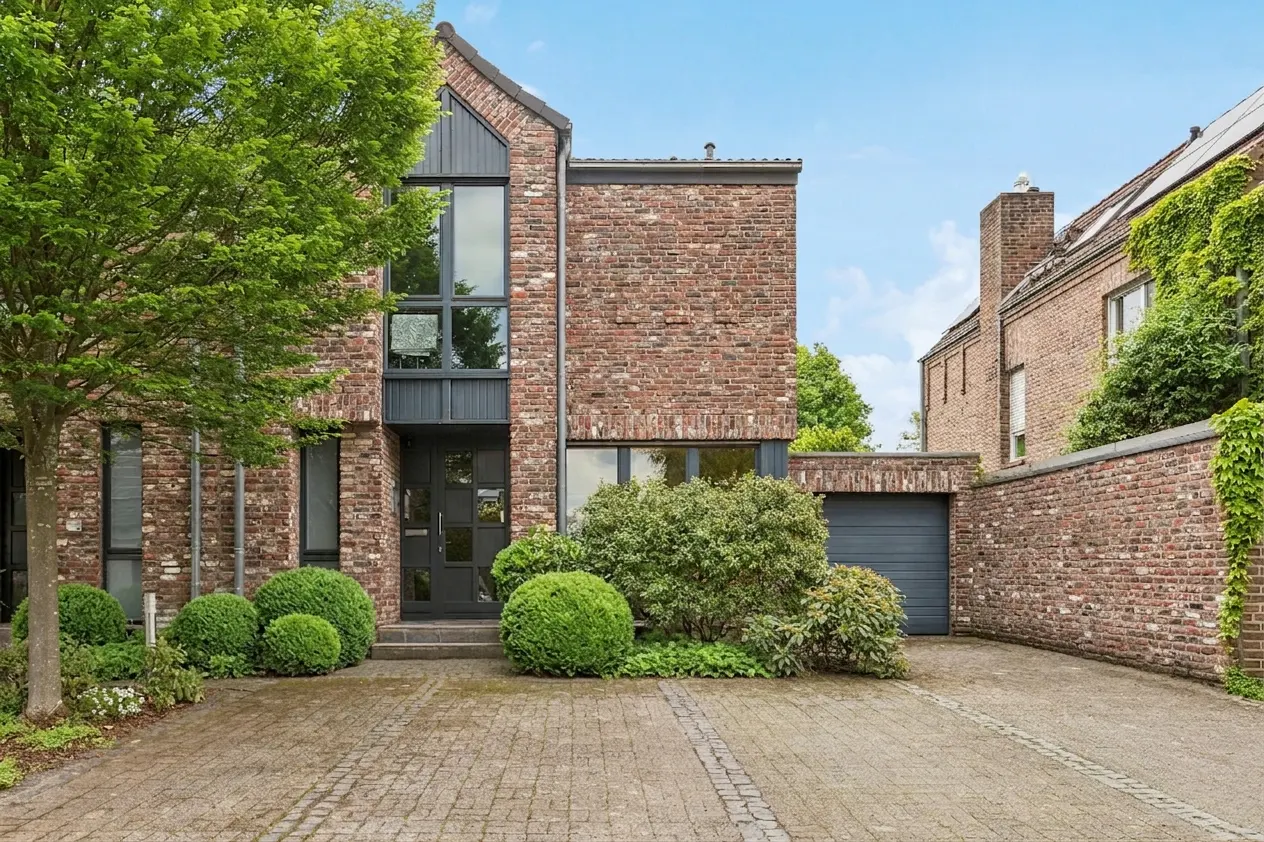 A two-story brick house with a gray door, windows, and garage door, surrounded by green bushes and a tree.