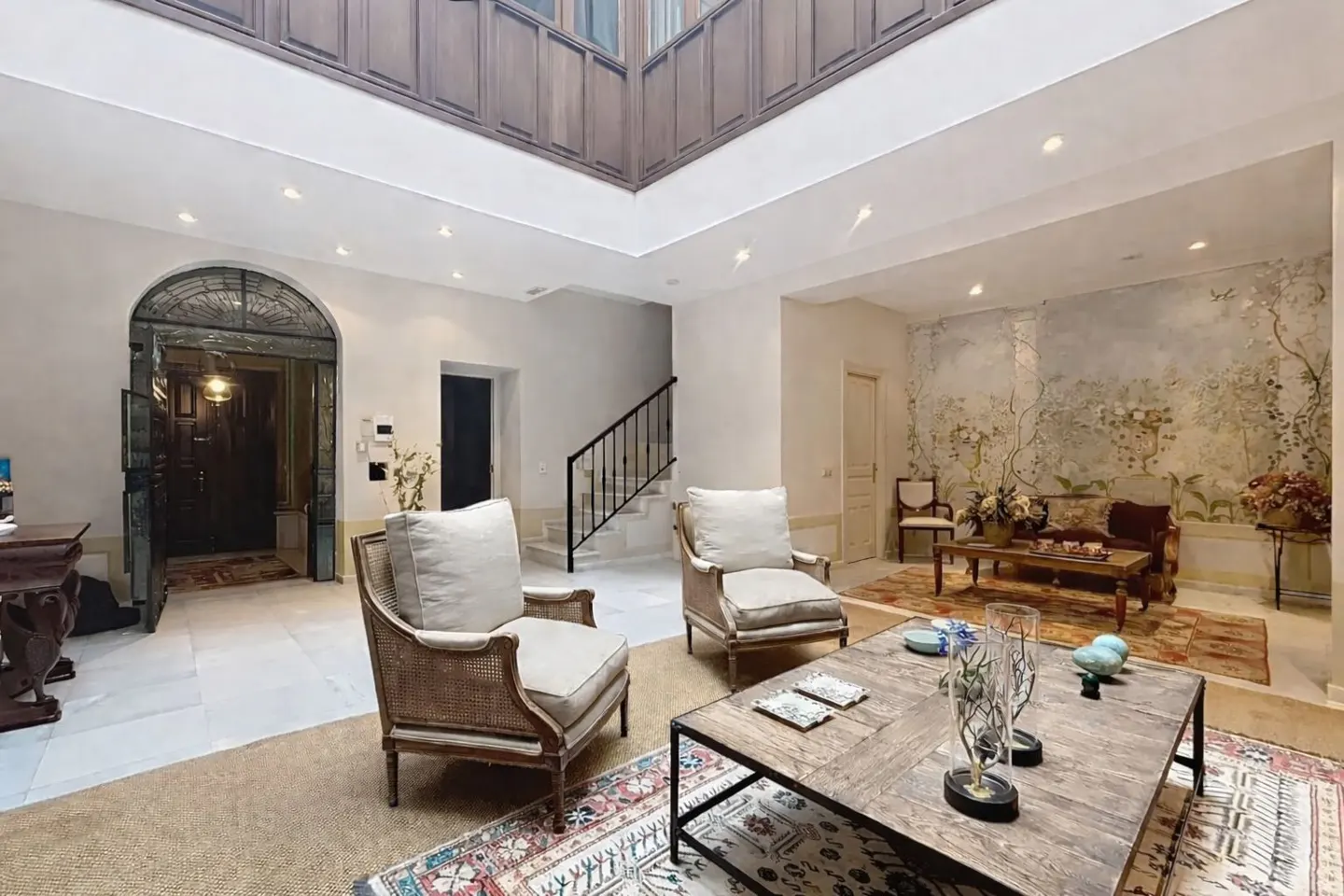 Bright living room with two chairs, a coffee table, and a floral mural. A staircase and arched doorway are visible.