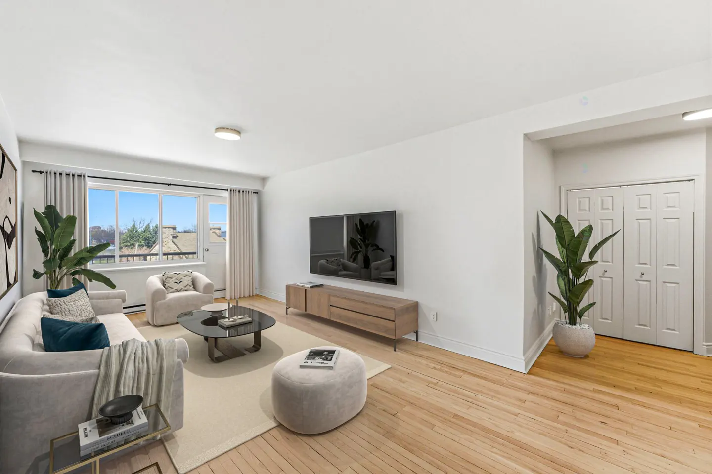 Bright living room with hardwood floors, white walls, and large window. Features a sofa, chair, TV, and plants.