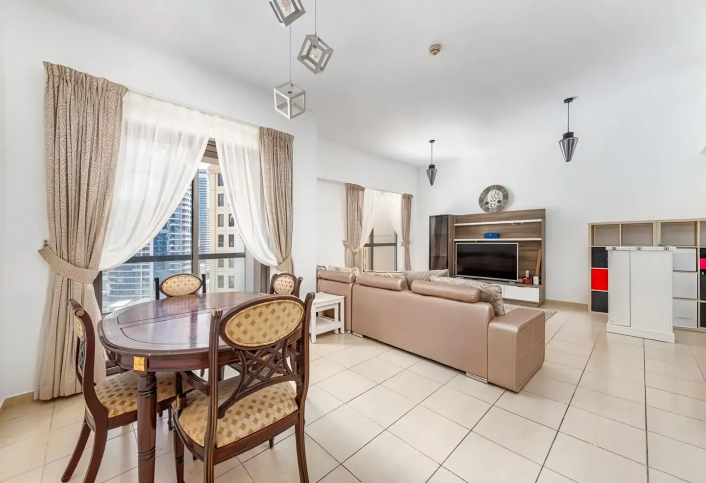 Bright living room with a dining set, beige leather sofa, TV unit, and large windows with city views and tiled floors.