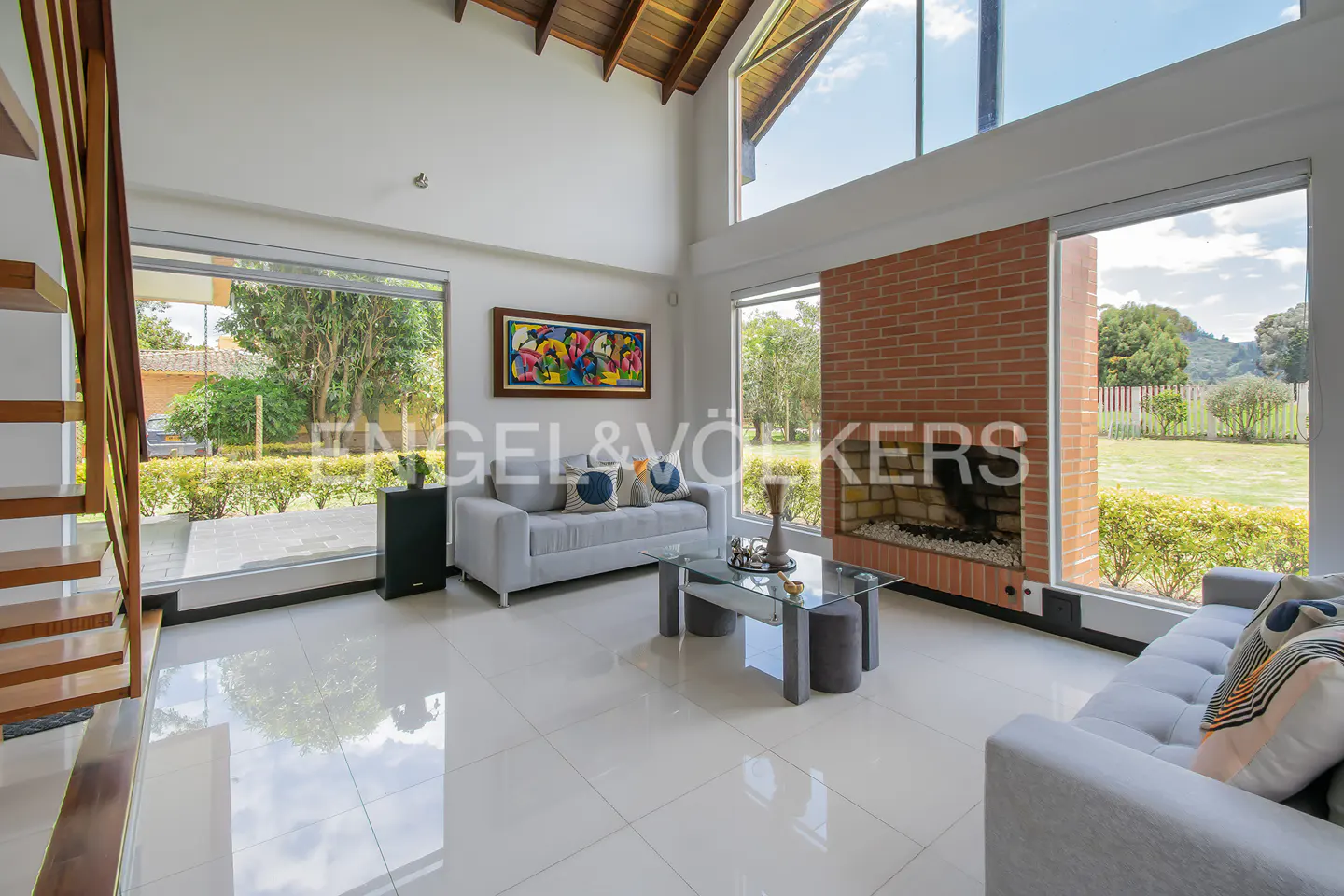 Bright living room with white tile floors, grey sofas, and a brick fireplace. Large windows offer views of the green landscape.
