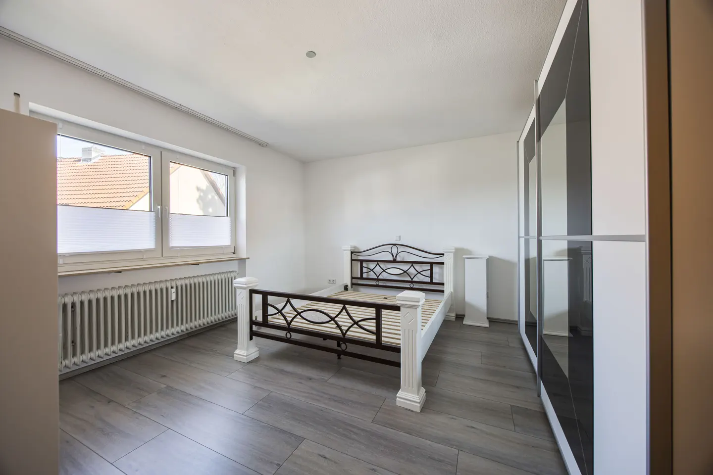 Bright bedroom with gray wood floors, white walls, and a black metal bed frame. A white wardrobe stands to the right.