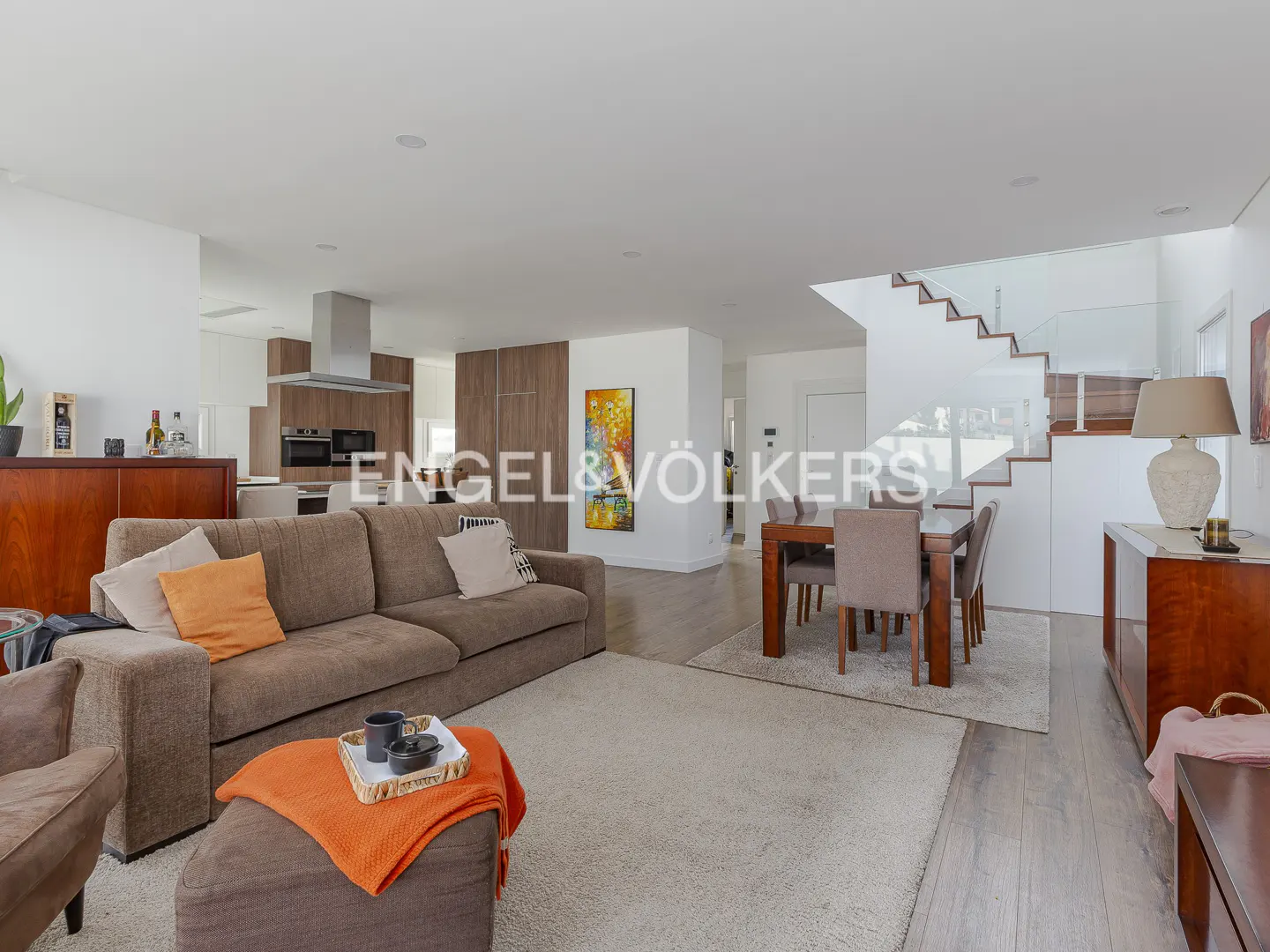Open-concept living space with a brown sofa, dining table, and modern staircase. The kitchen is visible in the background.