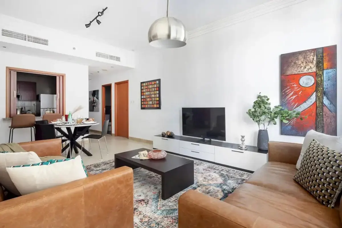 A bright living room with brown leather sofas, a dark wood coffee table, and a patterned rug. A dining area is visible through a window.