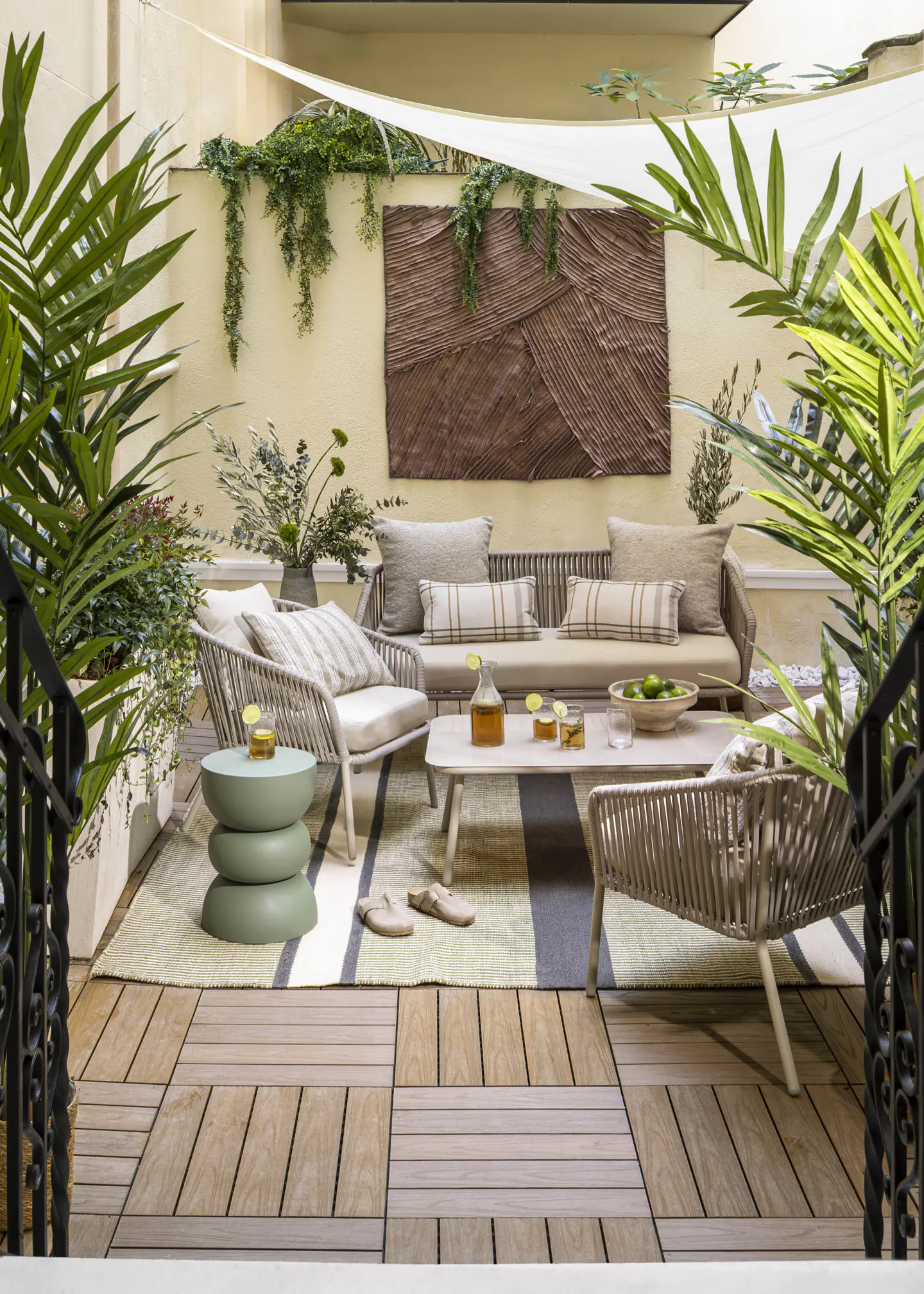 Outdoor patio with a sofa, chairs, and a coffee table on a striped rug. Green plants and a brown wall hanging add to the ambiance.