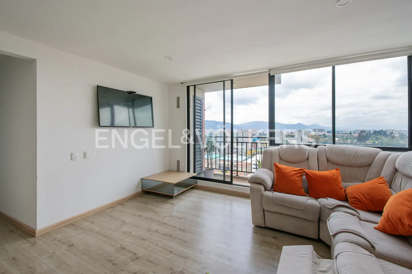 Bright living room with a beige sectional sofa, orange pillows, and a TV. A balcony offers city views. Engel & Völkers logo visible.