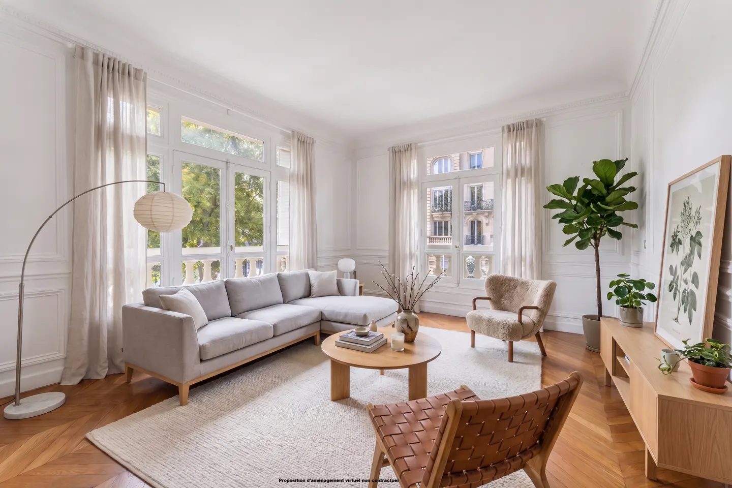 Bright living room with hardwood floors, white walls, and large windows. A gray sectional sofa, round wood table, and woven leather chair are featured.