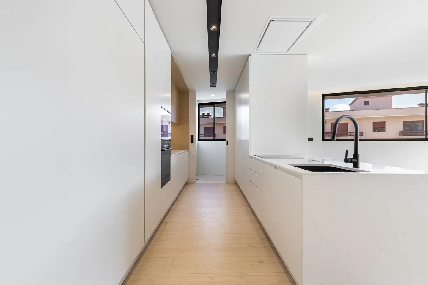 Modern minimalist kitchen with white cabinetry, light wood floors, black faucet, and a sleek linear ceiling light.
