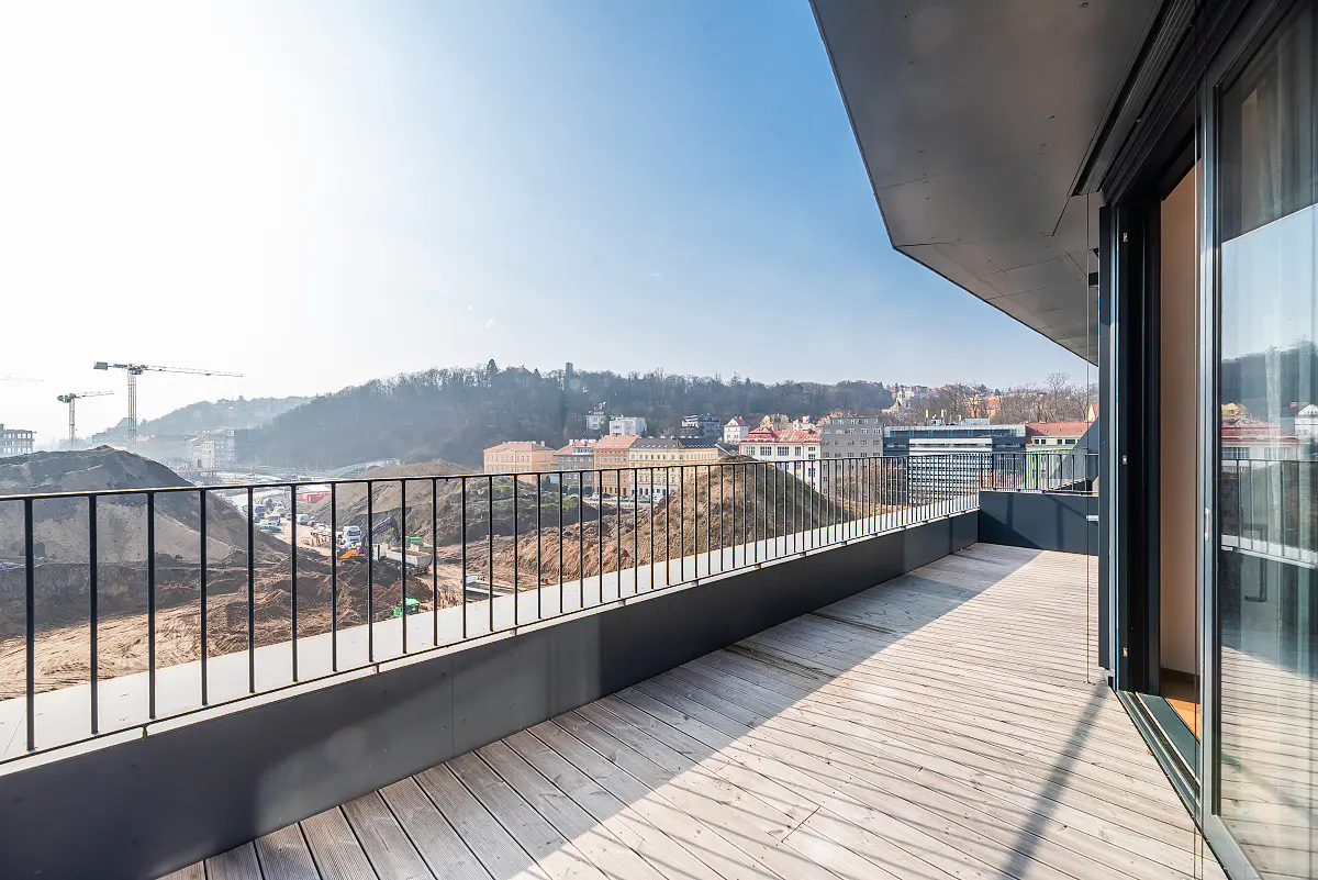 Balcony view with wood floor, black railing, and city view. Construction site with cranes visible in the background.