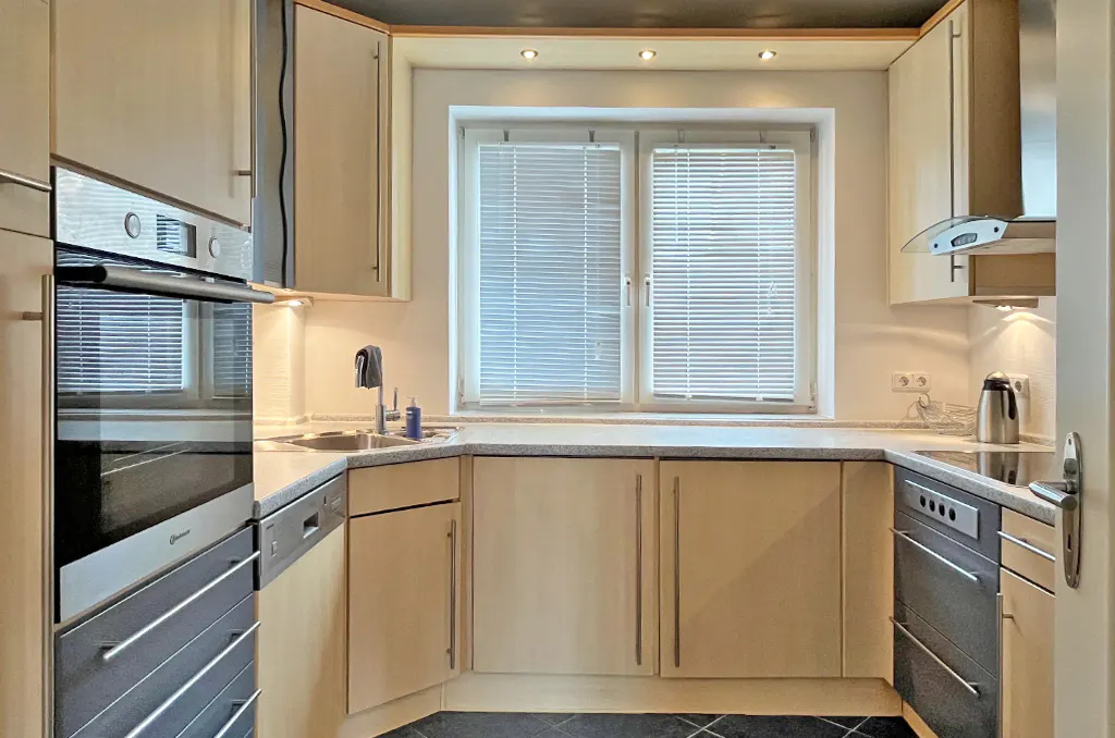 A bright kitchen with light wood cabinets, stainless steel appliances, and a window with blinds.