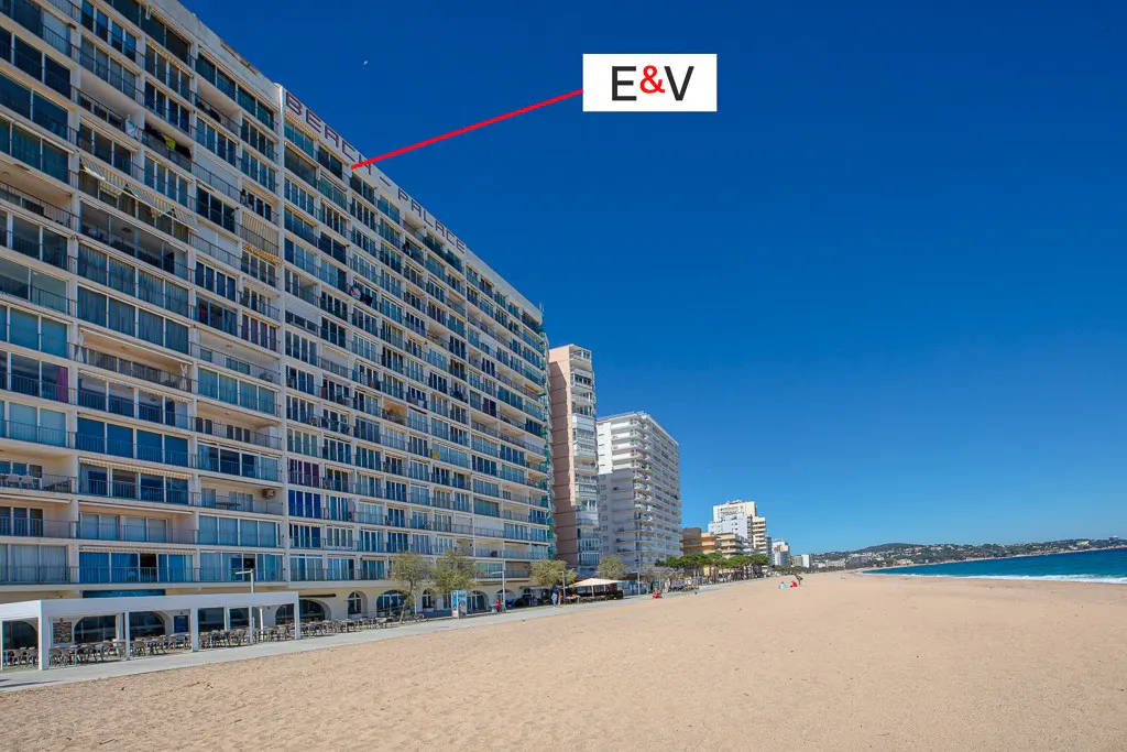Beachfront view of a tall, white apartment building with many balconies, next to a sandy beach and blue sky.