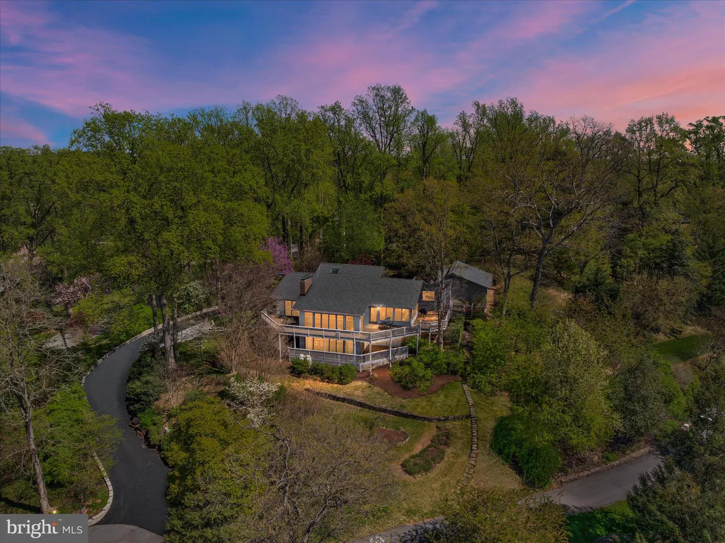 Aerial view of a modern gray house with a deck, surrounded by green trees under a pink and blue sky. A winding driveway leads to the house.