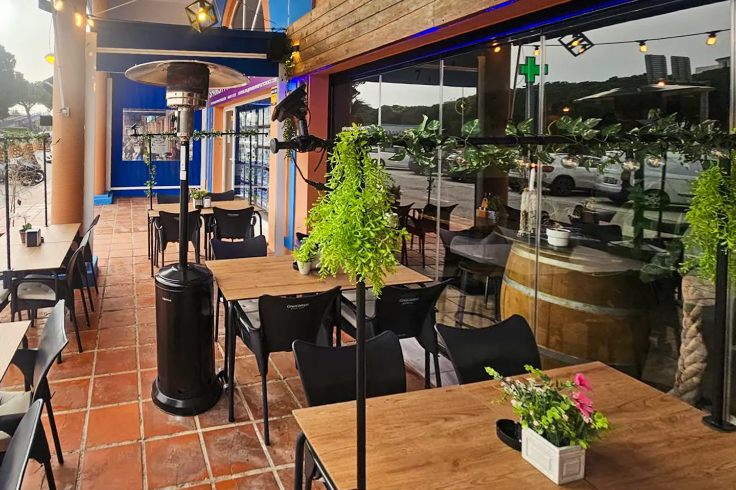 Outdoor restaurant patio with wooden tables, black chairs, and a black patio heater on a terracotta tiled floor. Green plants decorate the space.