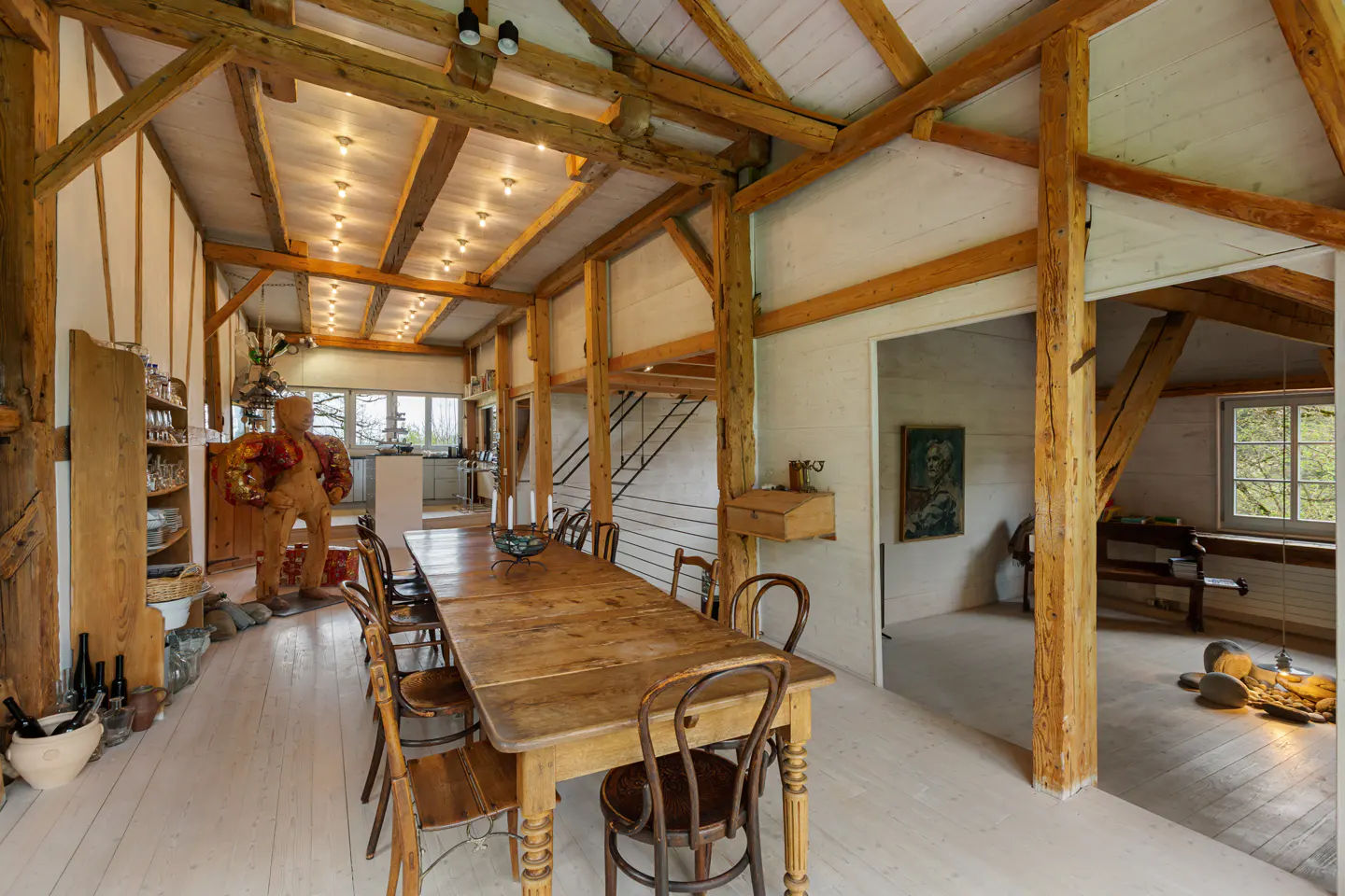 Rustic dining room with exposed wooden beams, white walls, and a long wooden table with chairs. A large sculpture stands near a shelf with glassware.