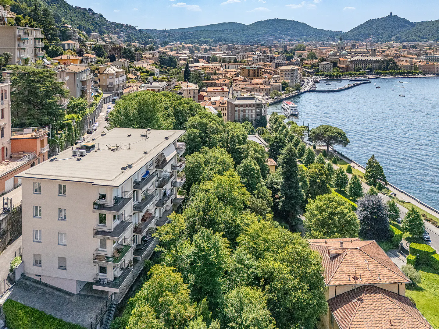 Aerial view of a multi-story apartment building surrounded by lush trees overlooking a lake and a coastal Italian town.