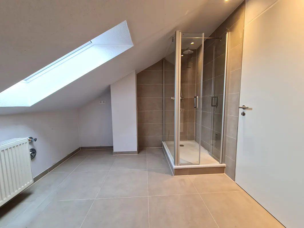 Attic bathroom with beige tile floor, white walls, skylight, radiator, and glass shower stall.