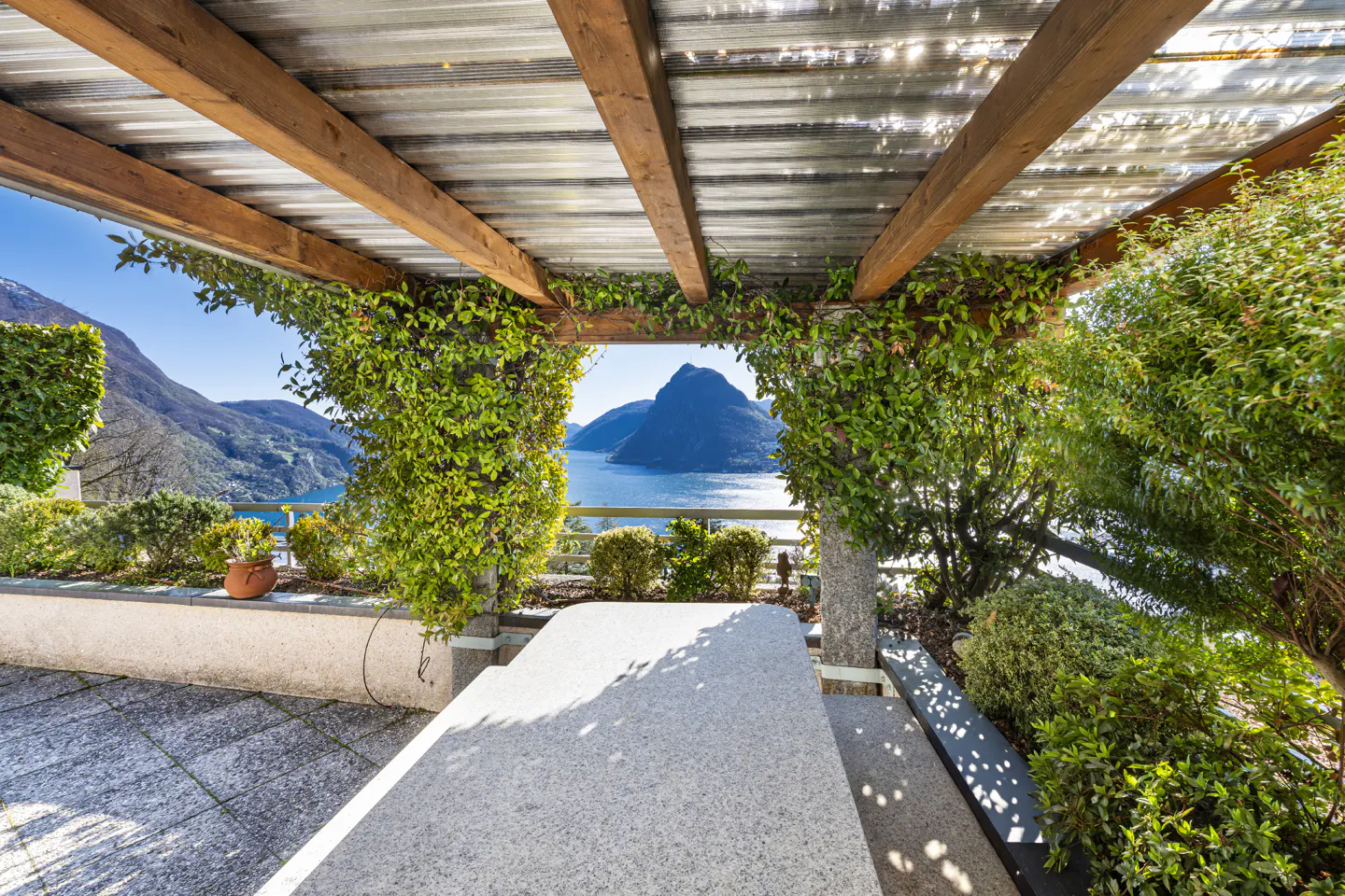 Outdoor patio with stone table and pergola covered in vines, overlooking a blue lake and mountains in the distance.