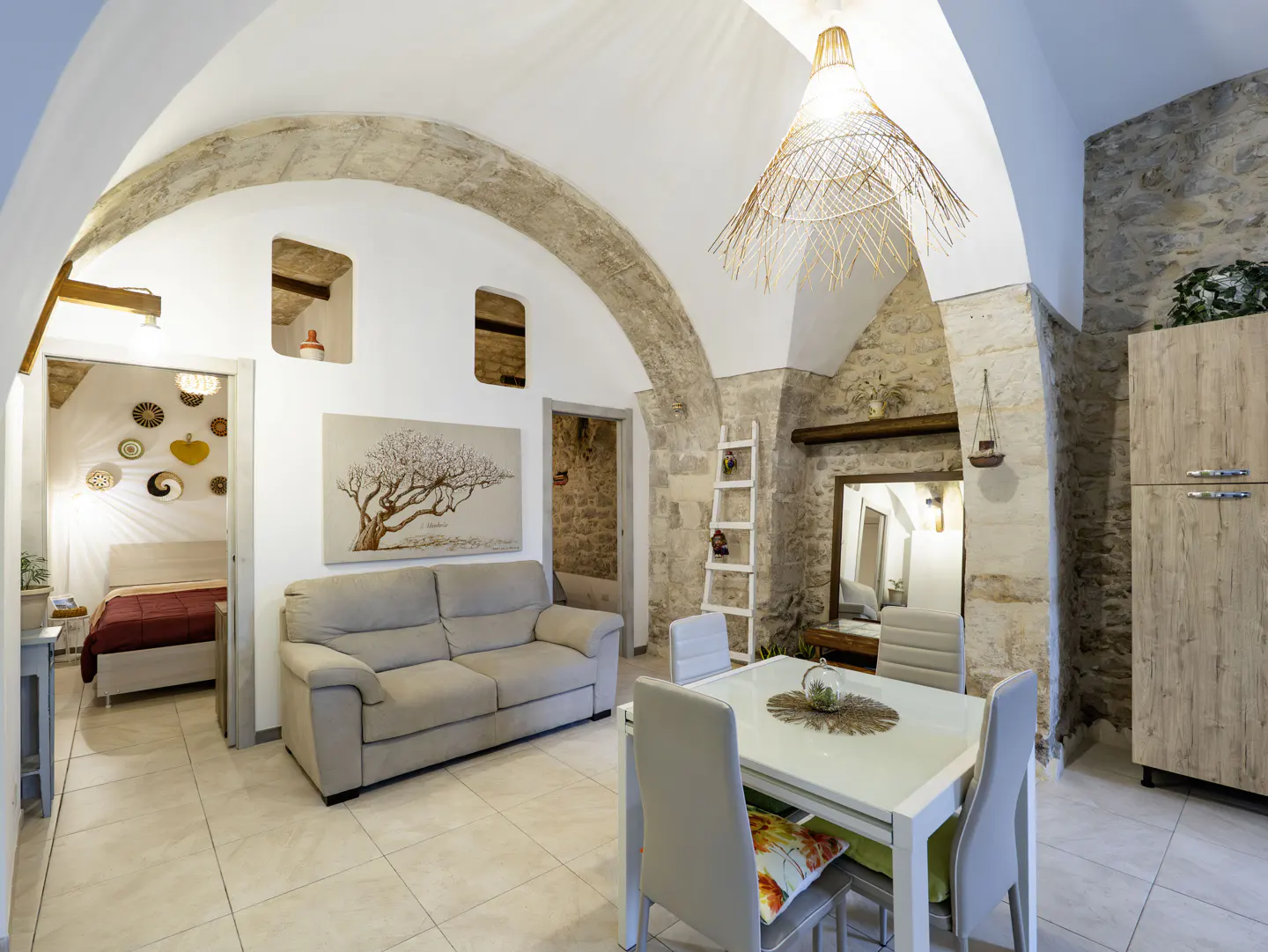 Bright living space with stone archways, a beige sofa, and a white dining table with four chairs. A painting of a tree hangs above the sofa.