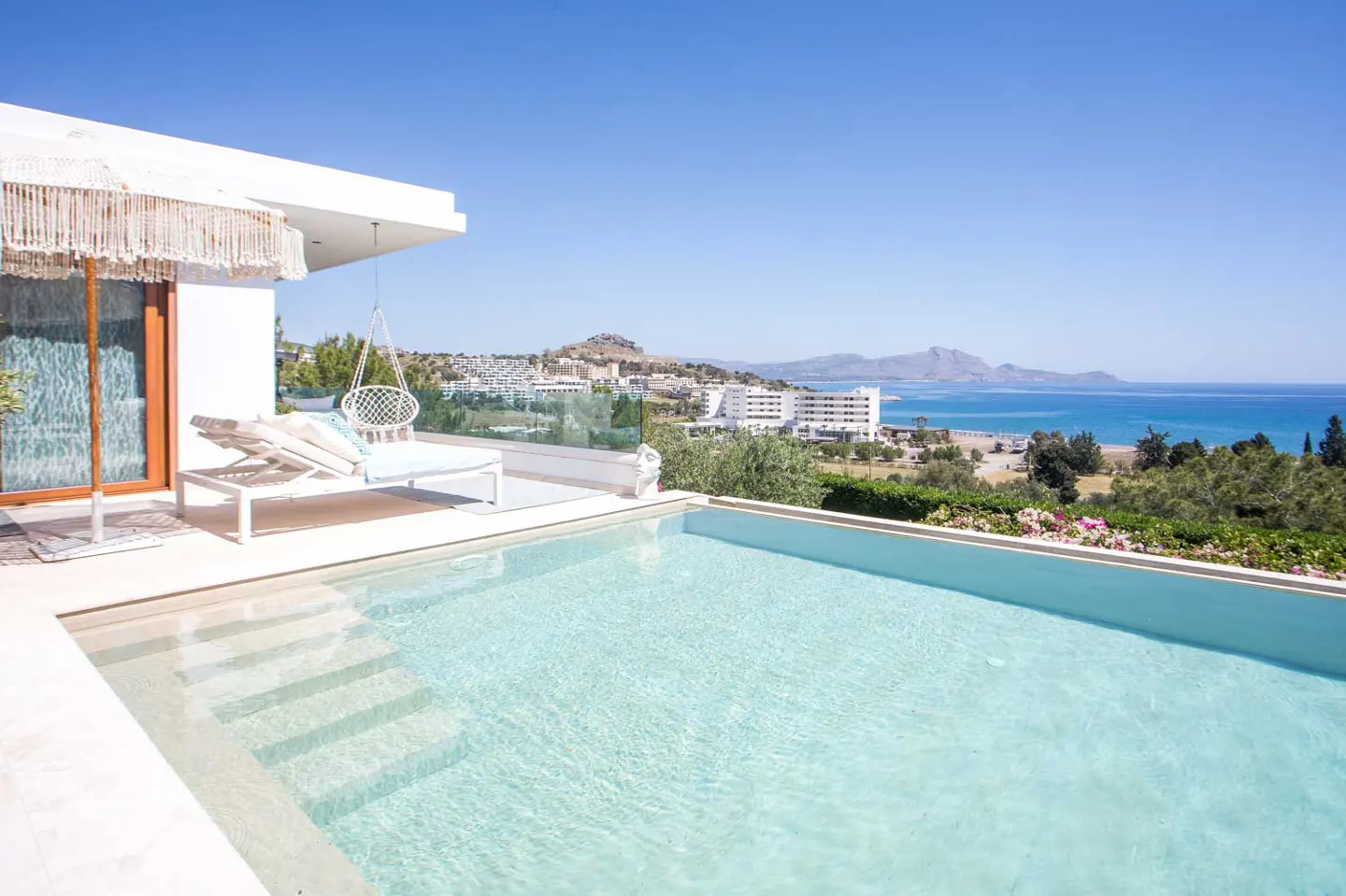 A modern white villa with a pool overlooking the ocean and a coastal town under a clear blue sky.