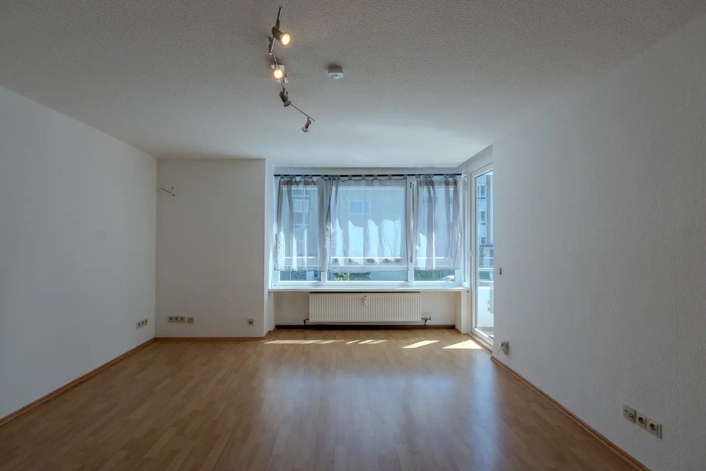 Empty room with white walls, light wood flooring, a large window with sheer curtains, and a modern track lighting fixture.