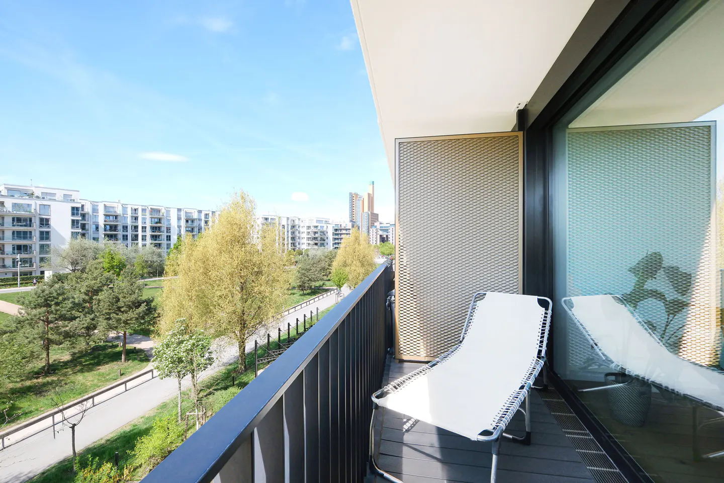 Balcony view with a white lounge chair, dark railing, and a view of a park with buildings in the background on a sunny day.