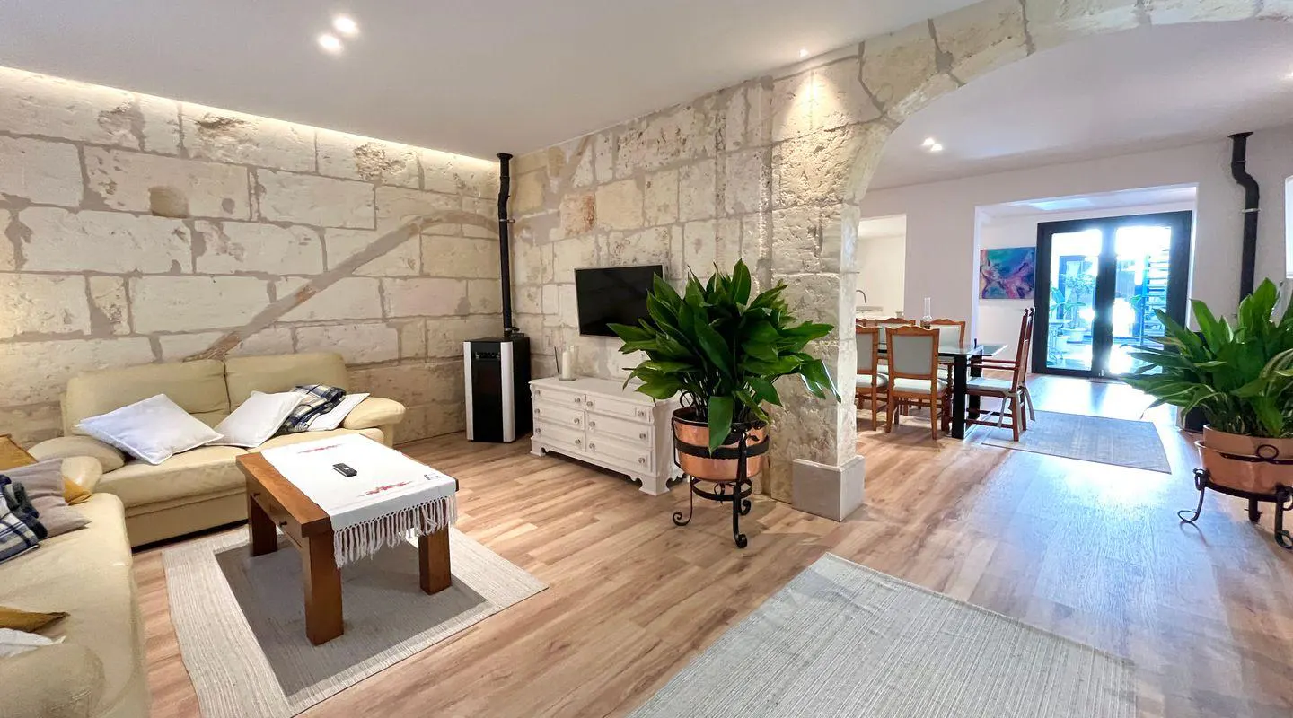 Living room with stone walls, wood floors, and an open dining area. A beige sofa, wood table, and potted plants add warmth to the space.