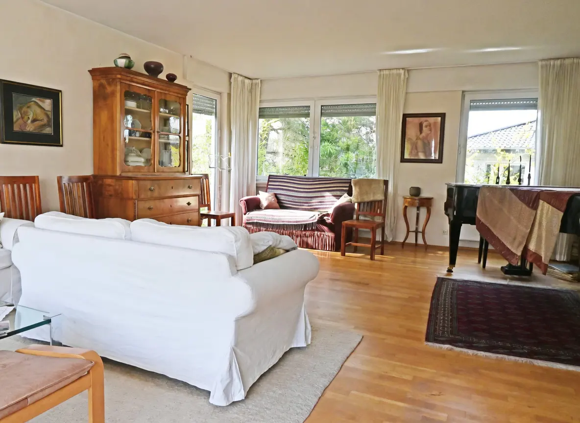 Bright living room with hardwood floors, white sofa, wooden hutch, striped couch, piano, and large windows.