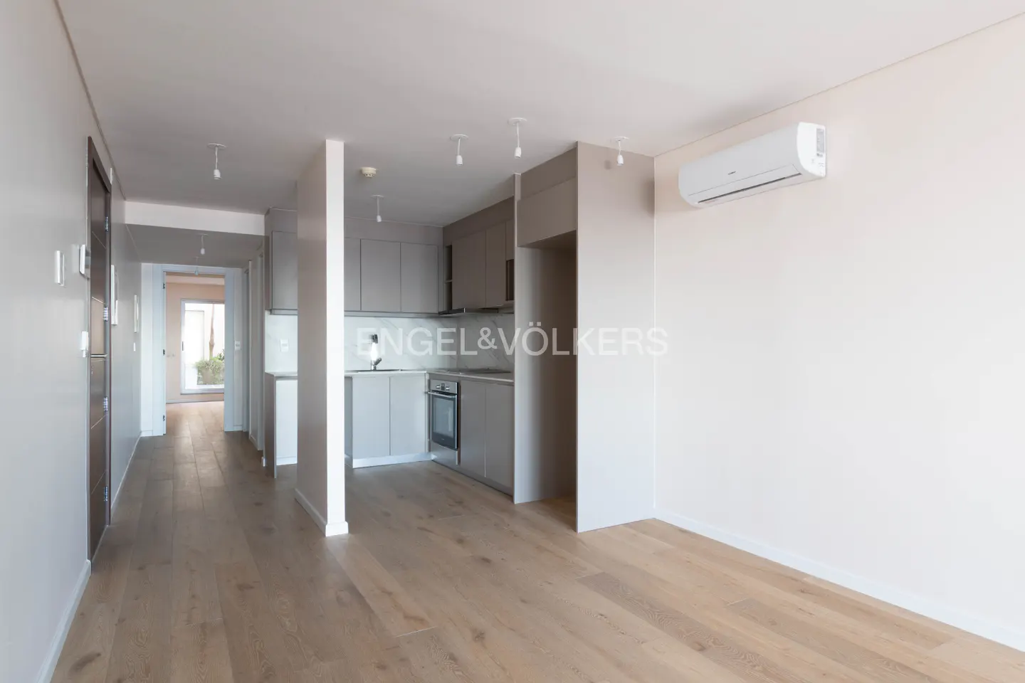 A bright, modern apartment interior with light wood floors, gray cabinets, and white walls. An air conditioner is mounted high on the wall.