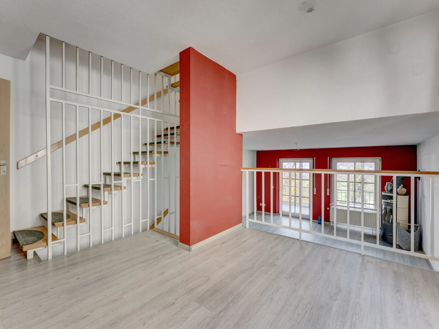 Interior view of a modern home with a staircase, red accent wall, and light wood floors. Windows provide natural light.