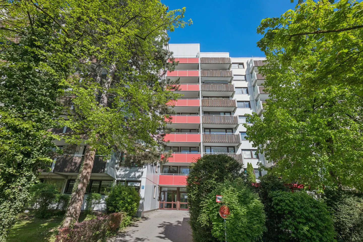 Exterior view of a tall, white apartment building with red balconies, framed by green trees under a blue sky.