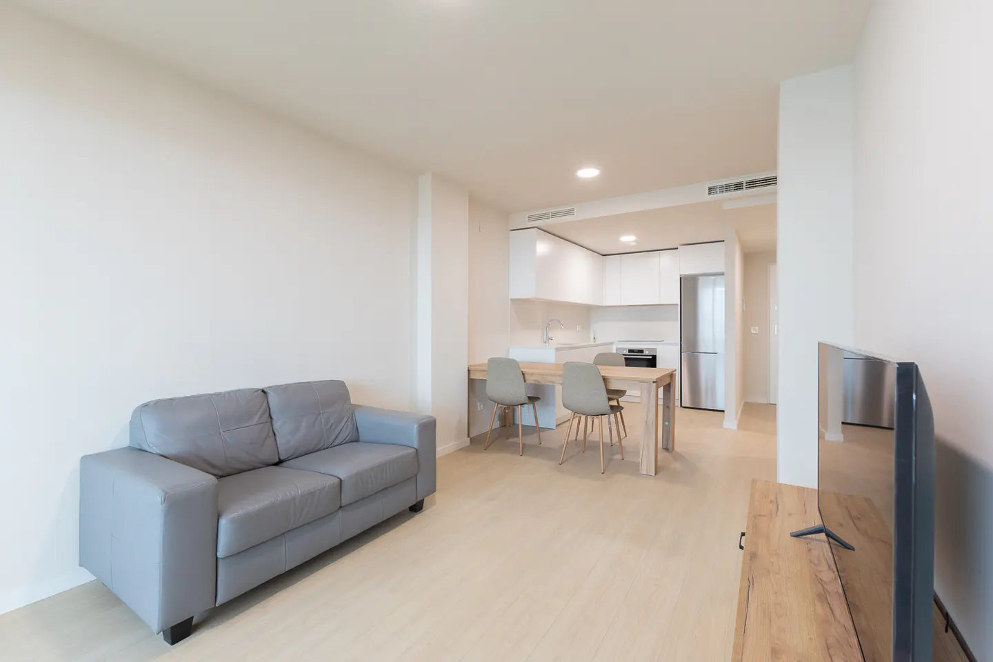 Bright, modern apartment with a gray sofa, wood table with chairs, and a white kitchen in the background. A TV is on the right.