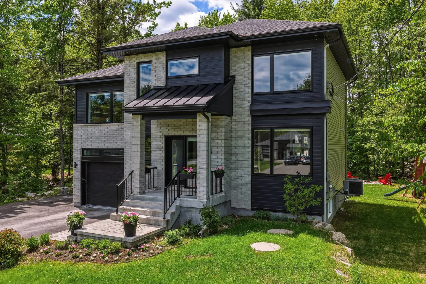 Modern two-story home with light brick and black siding, a front porch, and a lush green forest background.