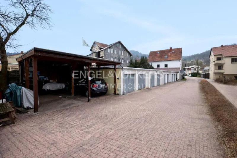 A paved driveway leads to a row of garages and a wooden carport with a parked car, set against a backdrop of houses.