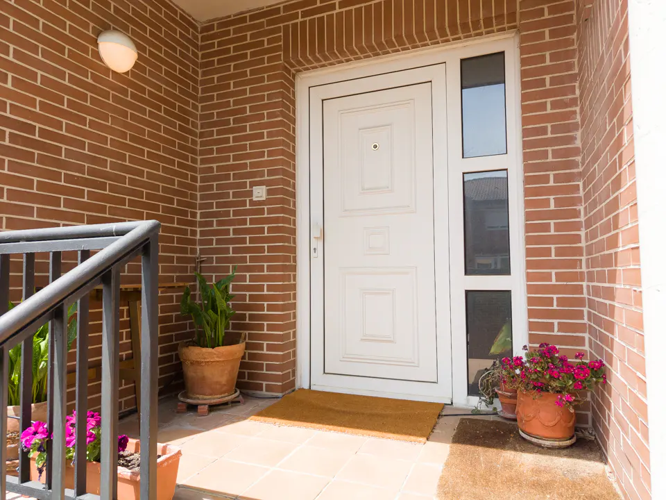 Front entrance of a brick house with a white door, sidelights, potted plants, and a brown doormat.