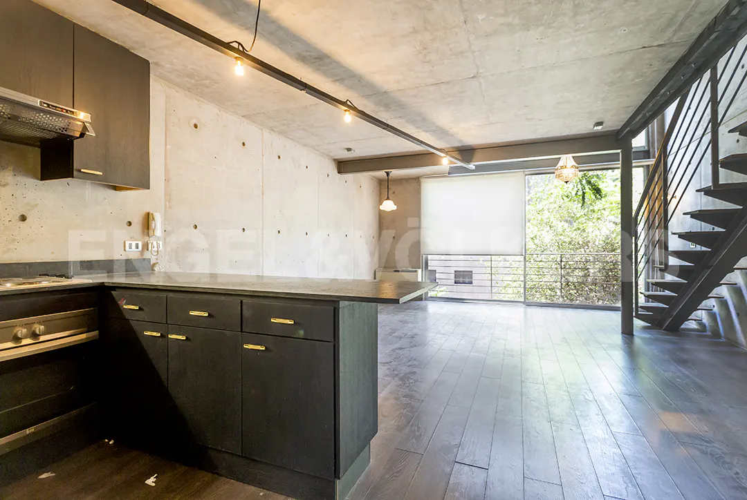 Modern apartment interior with dark wood floors, concrete walls, and black kitchen cabinets with gold handles. Metal staircase and large windows.