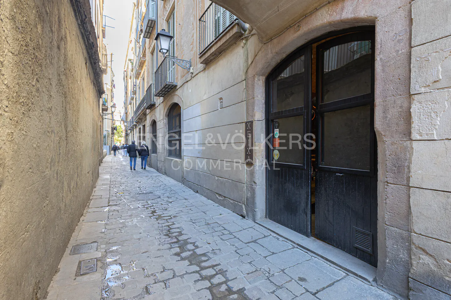 Narrow cobblestone street with people walking away. Buildings with balconies line the street. Black door on the right.